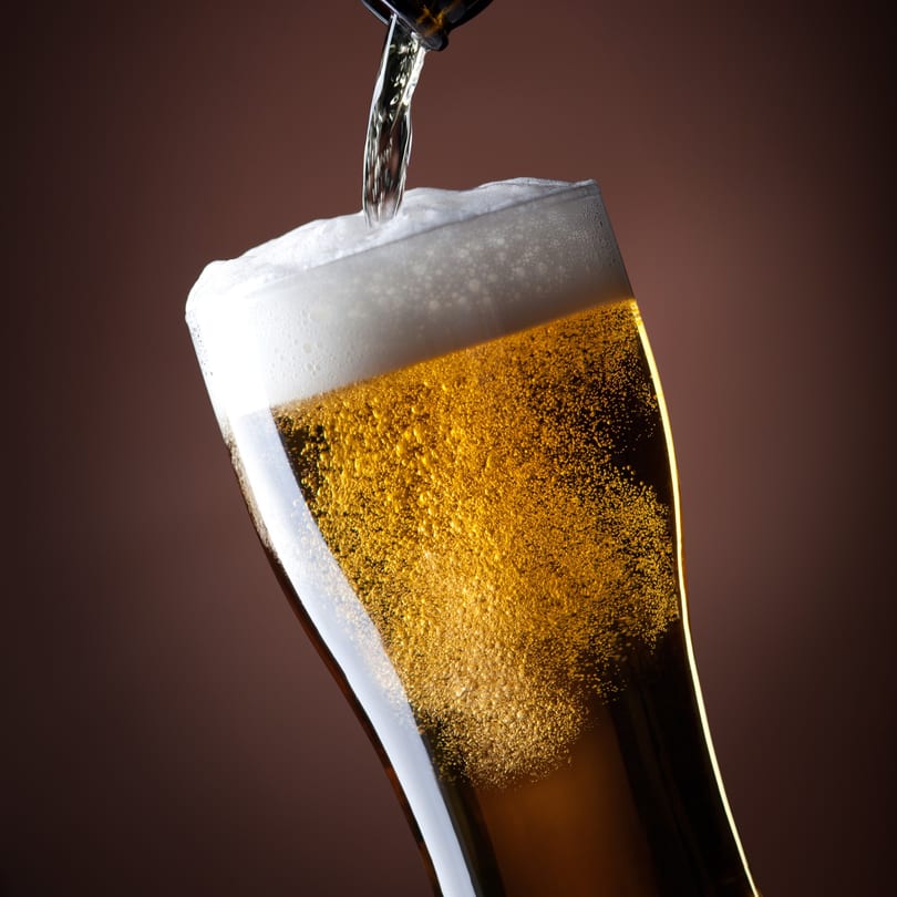 Golden beer with a frothy head being poured into a glass against a dark background.