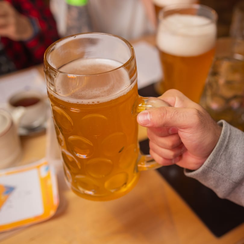 A hand holding a large glass mug filled with golden beer, with other drinks blurred in the background.
